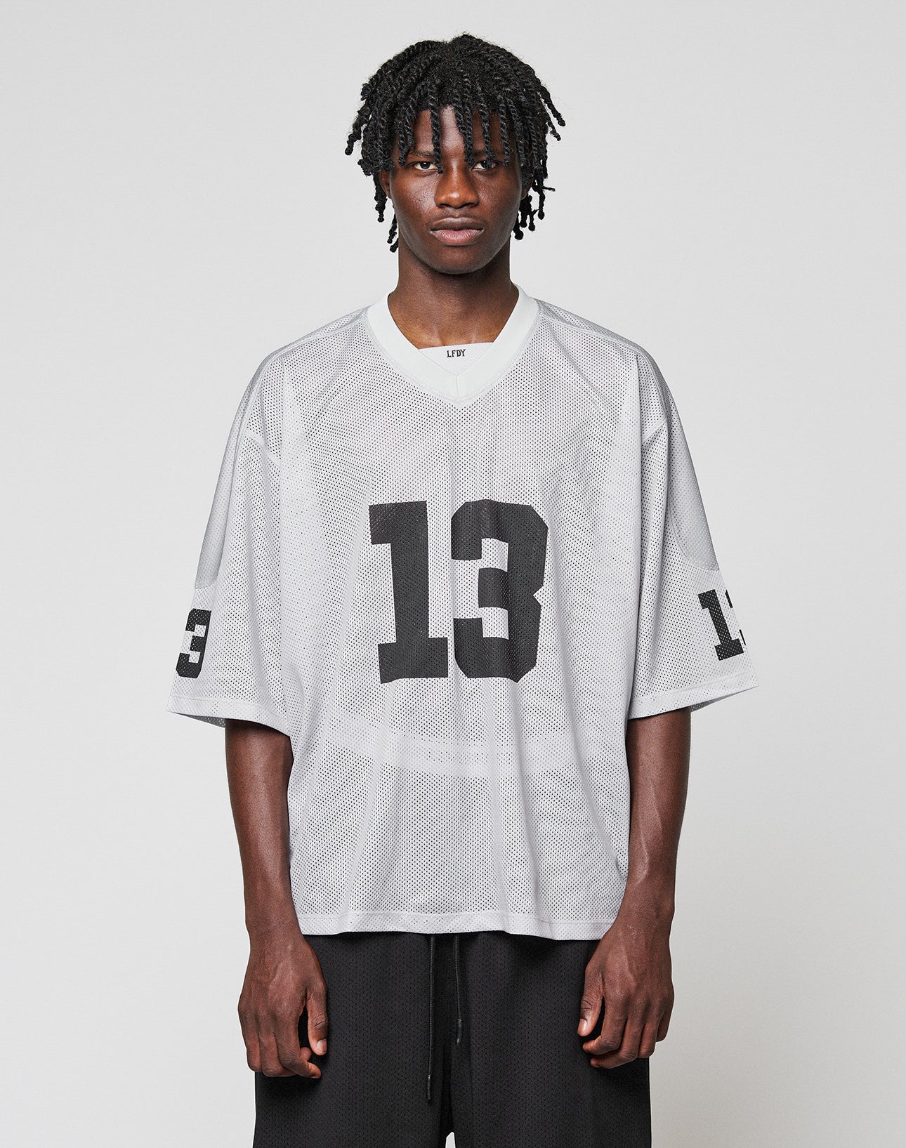 A man with short dreadlocks wears the LIVE FAST DIE YOUNG LFDY Mesh Jersey—an oversized, light gray polyester jersey with a V-neck and black number 13 on the front and sleeves—against a plain light background. | model