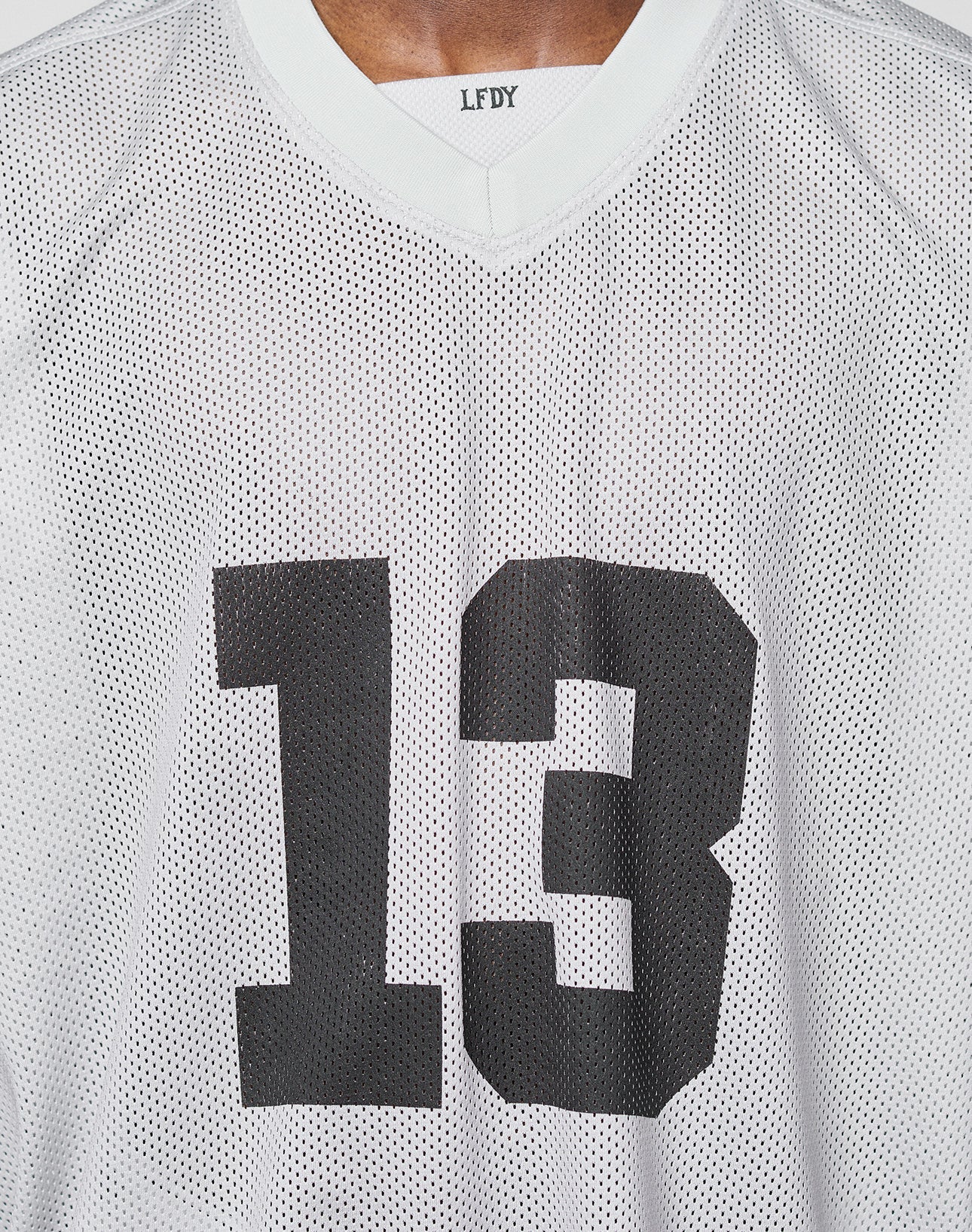 A close-up of a person wearing the LFDY Mesh Jersey by LIVE FAST DIE YOUNG, featuring a white polyester mesh, black number 13 on the front, V-neck design, and LFDY lettering inside the collar.