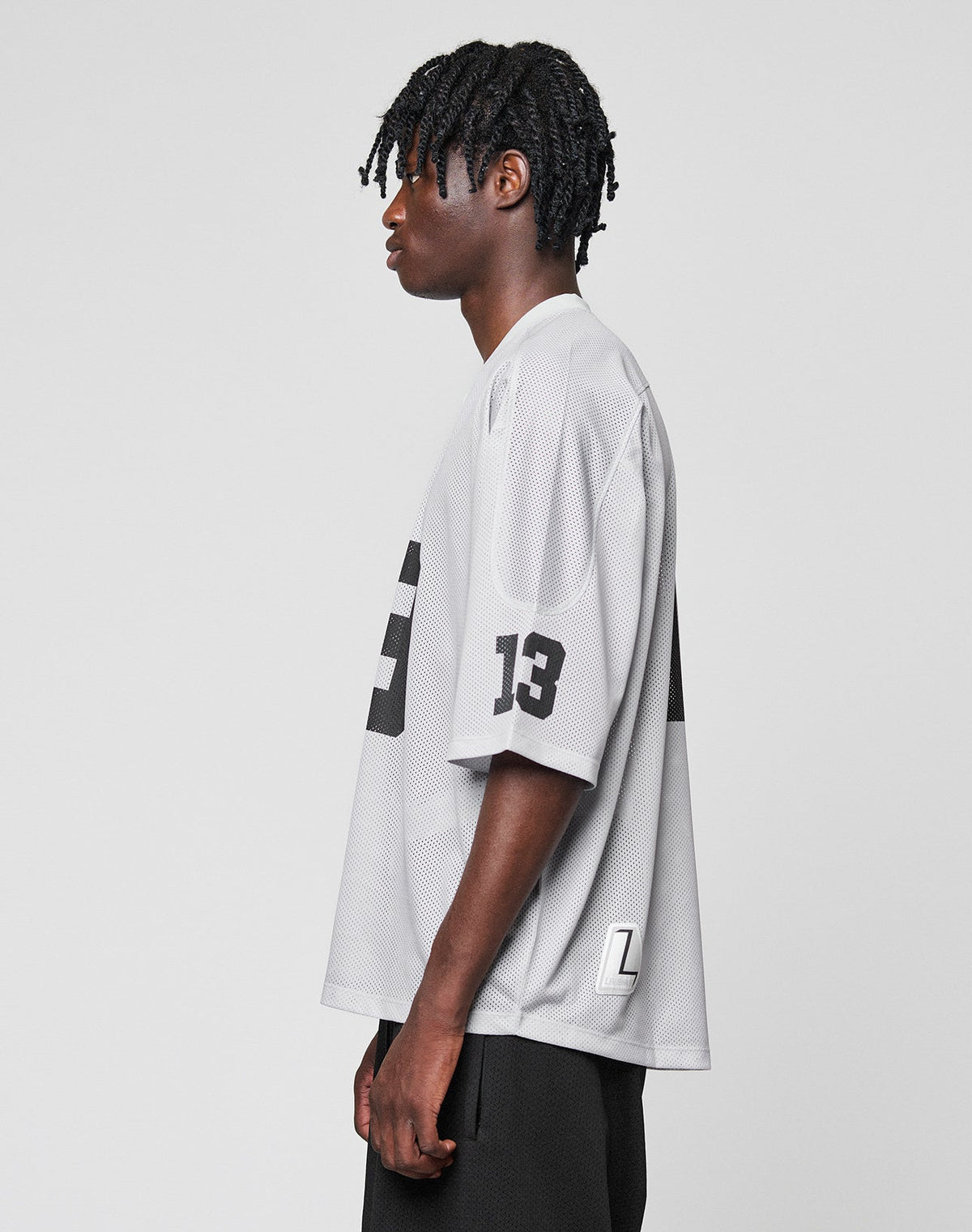 A young man with short dreadlocks stands in profile wearing the LIVE FAST DIE YOUNG LFDY Mesh Jersey, a loose-fitting, light gray polyester sports top with black number 13 details and V-neck, paired with black shorts against a plain background.
