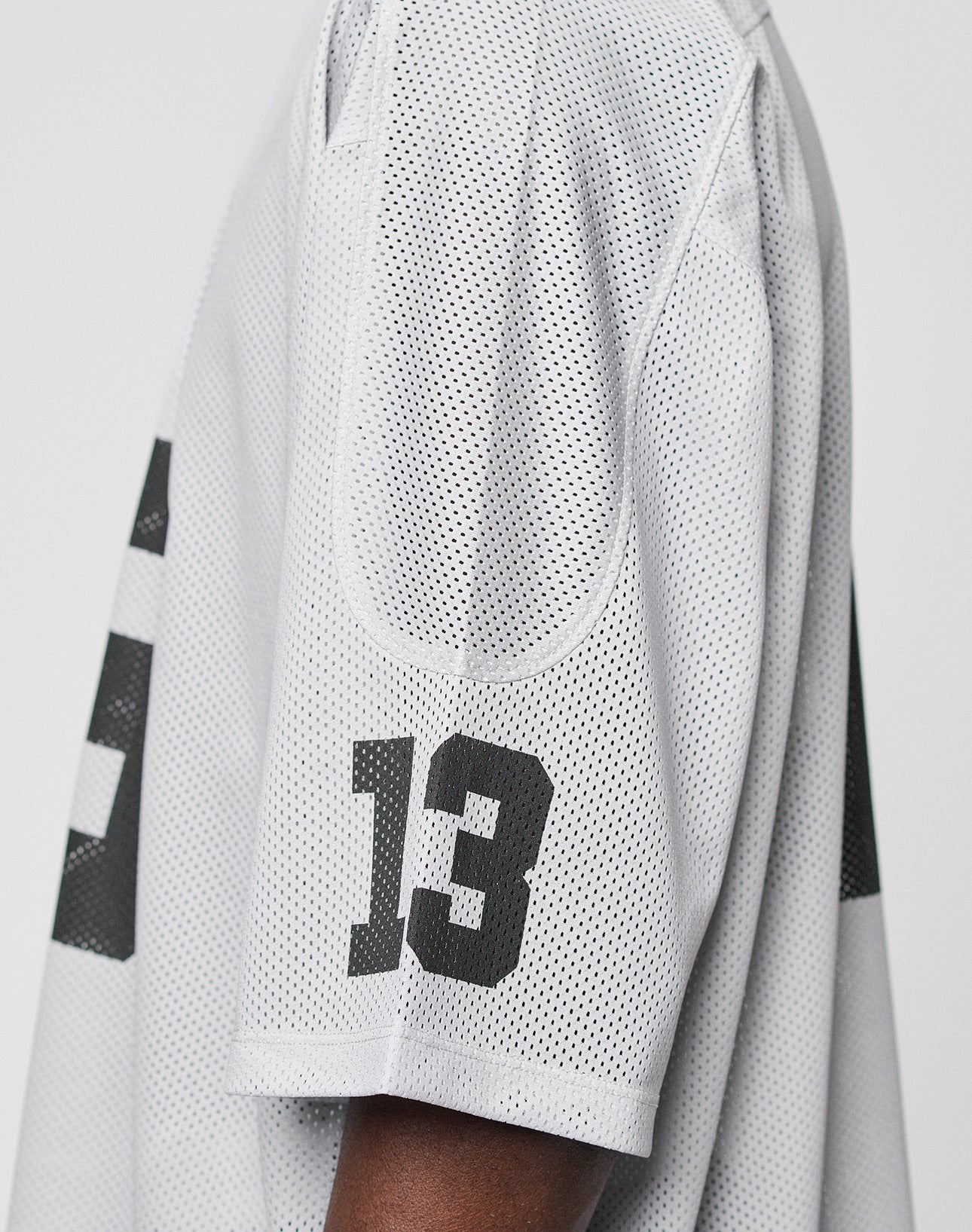 Close-up of a persons arm in the LFDY Mesh Jersey by LIVE FAST DIE YOUNG, featuring black number 13 on the sleeve. The oversize white polyester mesh design offers comfort against a plain, light background.