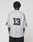 A person with short twisted hair stands with their back to the camera, wearing the oversize-fit white LFDY Mesh Jersey by LIVE FAST DIE YOUNG featuring a V-neck and black number 13, paired with black shorts.