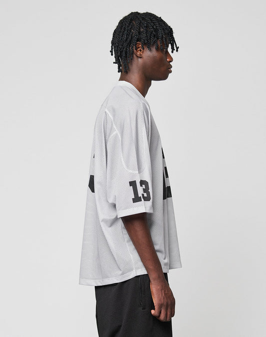 A young man with short twists stands in profile against a white background, wearing the LIVE FAST DIE YOUNG LFDY Mesh Jersey in light gray with an oversize fit, mesh fabric, number 13 on the sleeve, and a V-neck.