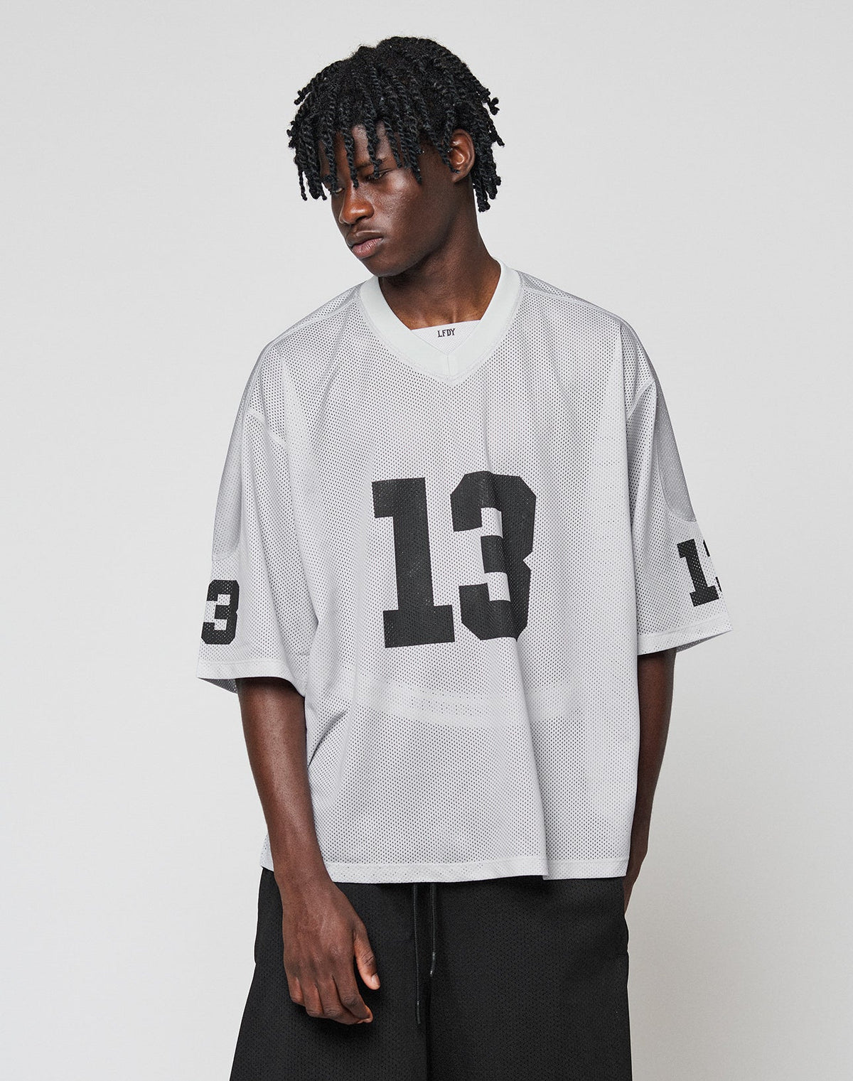 A person with short, textured hair models the oversized LFDY Mesh Jersey by LIVE FAST DIE YOUNG in gray polyester mesh with black number 13 on the front and sleeve, paired with black pants against a plain background.