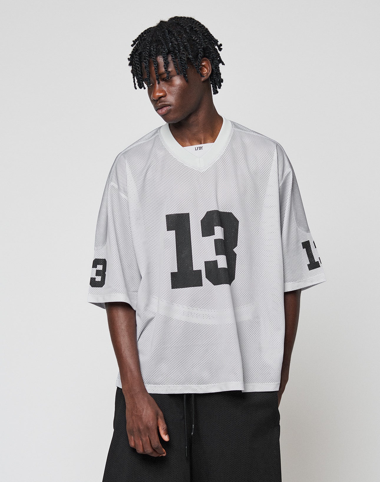 A person with short, textured hair models the oversized LFDY Mesh Jersey by LIVE FAST DIE YOUNG in gray polyester mesh with black number 13 on the front and sleeve, paired with black pants against a plain background.
