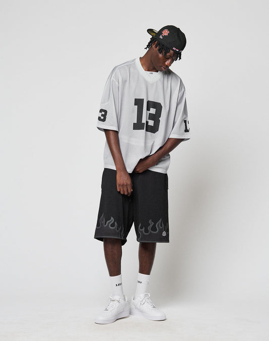 A person in a black cap, LIVE FAST DIE YOUNG LFDY Mesh Jersey (gray, number 13, v-neck), black flame shorts, white socks, and sneakers stands against a white background looking down.