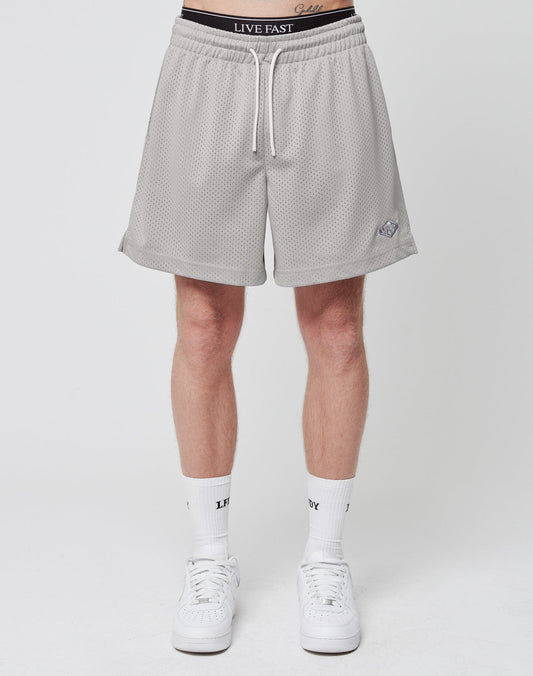 A person stands against a plain background in LIVE FAST DIE YOUNG Basic Mesh Shorts, light gray with a logo patch and white drawstrings at the waist. Made from 100% polyester, theyre styled with white socks and sneakers; LIVE FAST is visible on waistband and socks.