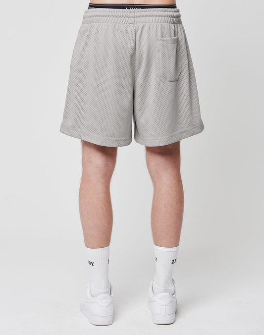 A person faces away, wearing LIVE FAST DIE YOUNG Basic Mesh Shorts in light gray (100% polyester, regular fit), featuring a logo patch, elastic waistband, and small back pocket. Paired with white crew socks and sneakers. Plain white background.