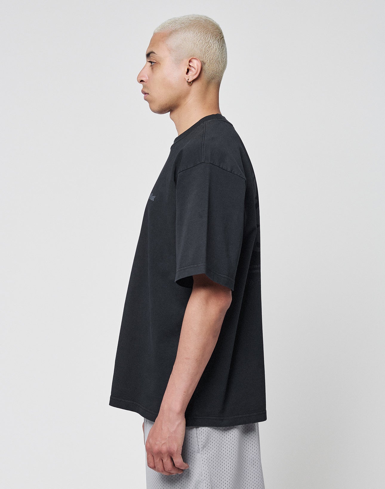 A person with short, bleached blond hair stands in profile against a plain background, wearing a loose-fitting LIVE FAST DIE YOUNG Basic 280 Tee in Oversize Fit and light-colored shorts.