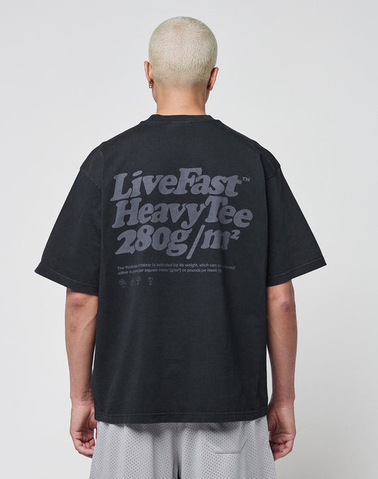 A person with short, bleached blonde hair is shown from behind, wearing a black LIVE FAST DIE YOUNG Basic 280 Tee with a bold backprint. The background is plain white.