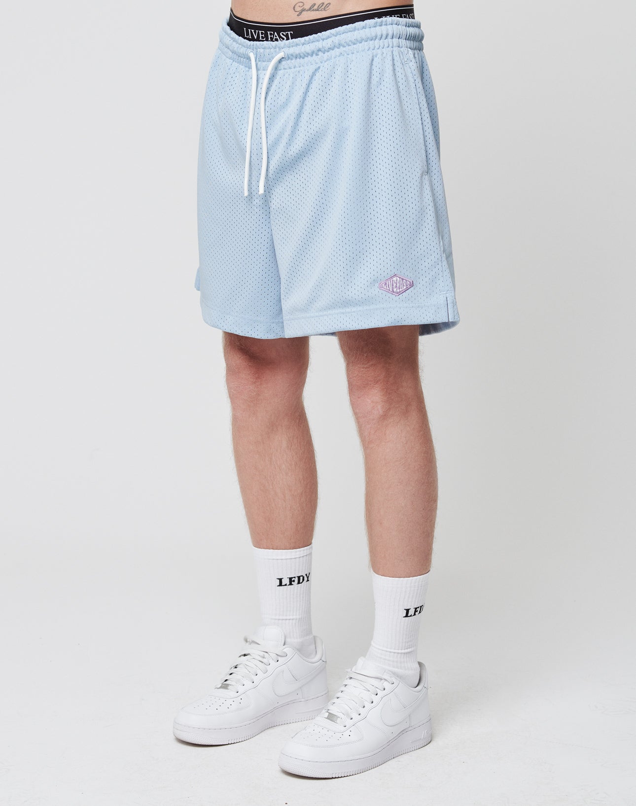 A person stands against a plain background wearing LIVE FAST DIE YOUNG Basic Mesh Shorts in light blue, white crew socks with black text, and white Nike sneakers. Only the lower half of the body is visible.