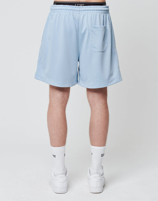 A person stands facing away, wearing LIVE FAST DIE YOUNG Basic Mesh Shorts in light blue. These regular fit, 100% polyester shorts have an elastic waistband and a back pocket. Only the lower body is visible against a plain white background.