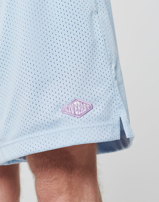Close-up of a persons upper leg wearing LIVE FAST DIE YOUNG Basic Mesh Shorts in light blue. These regular fit polyester shorts feature an elastic waistband, side slit for comfort, and a small lavender diamond-shaped GIVELAST patch near the hem.