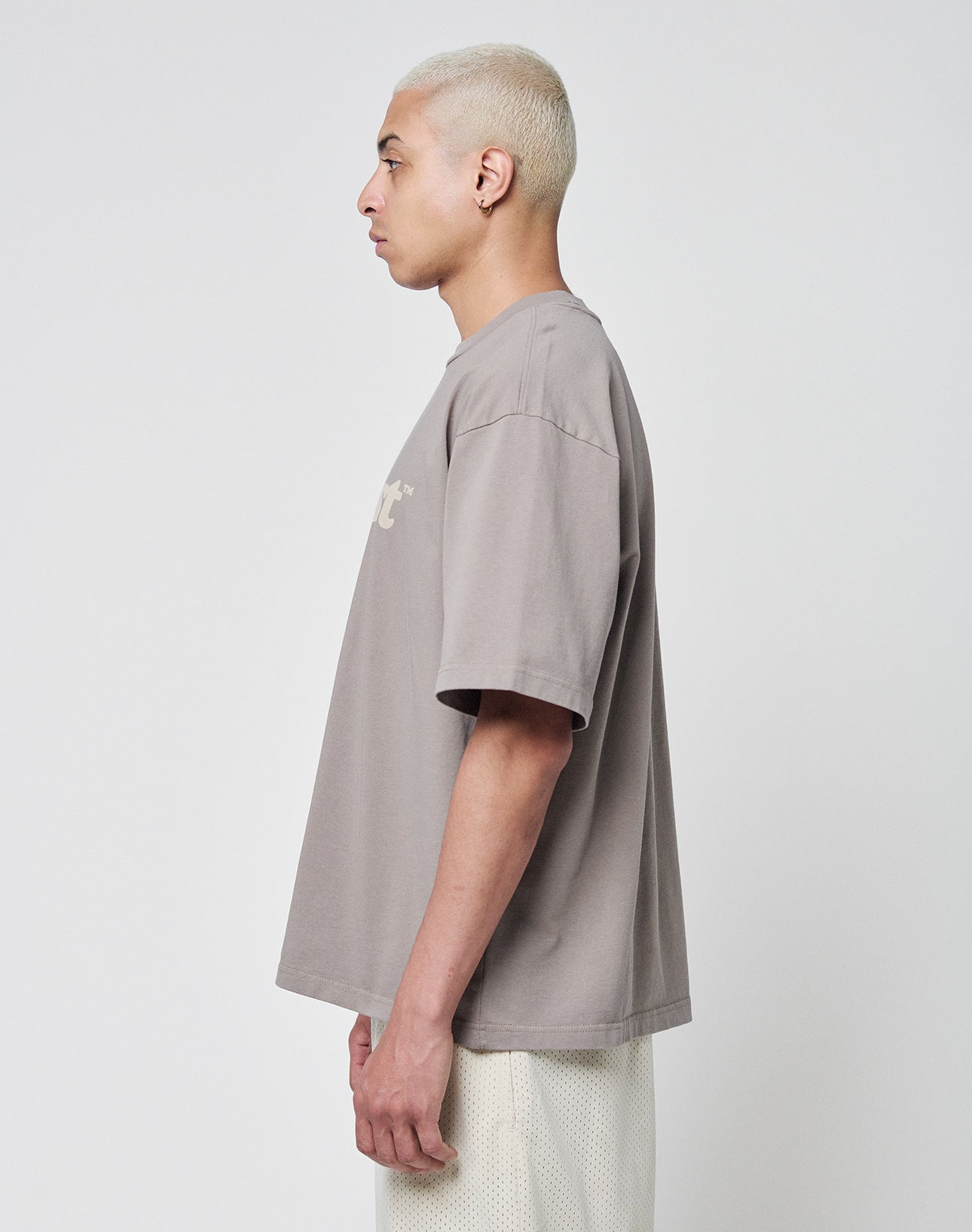 A person with short, bleached blond hair stands in profile against a plain background, wearing the LIVE FAST DIE YOUNG Basic Tee in an oversized fit and white mesh shorts.