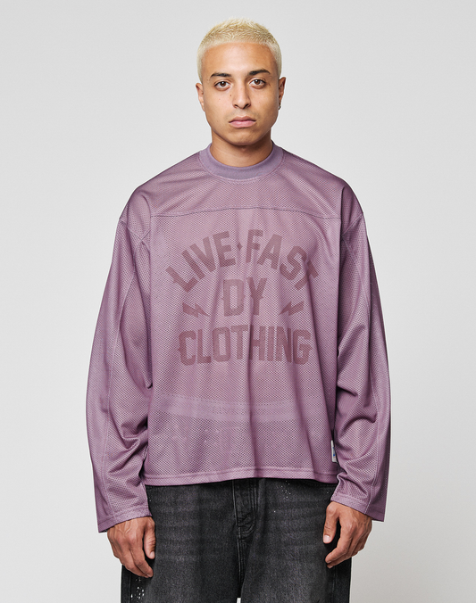 A person with short, bleached blond hair faces the camera wearing the LFDY Flash Jersey by LIVE FAST DIE YOUNG—an oversized purple mesh long-sleeve with a bold front print—paired with black jeans against a light gray background. | model