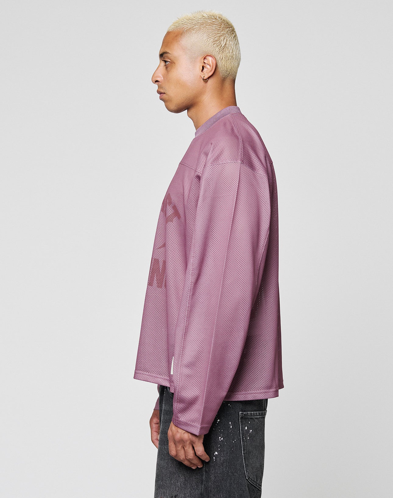 A person with short, bleached blond hair stands in profile against a plain light gray background, wearing an oversize fit LFDY Flash Jersey by LIVE FAST DIE YOUNG and dark, paint-splattered jeans.