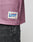 Close-up of a pink LFDY Flash Jersey by LIVE FAST DIE YOUNG with an oversize fit and a rectangular patch label with text and an M for size, above faded black jeans with white paint splatters.