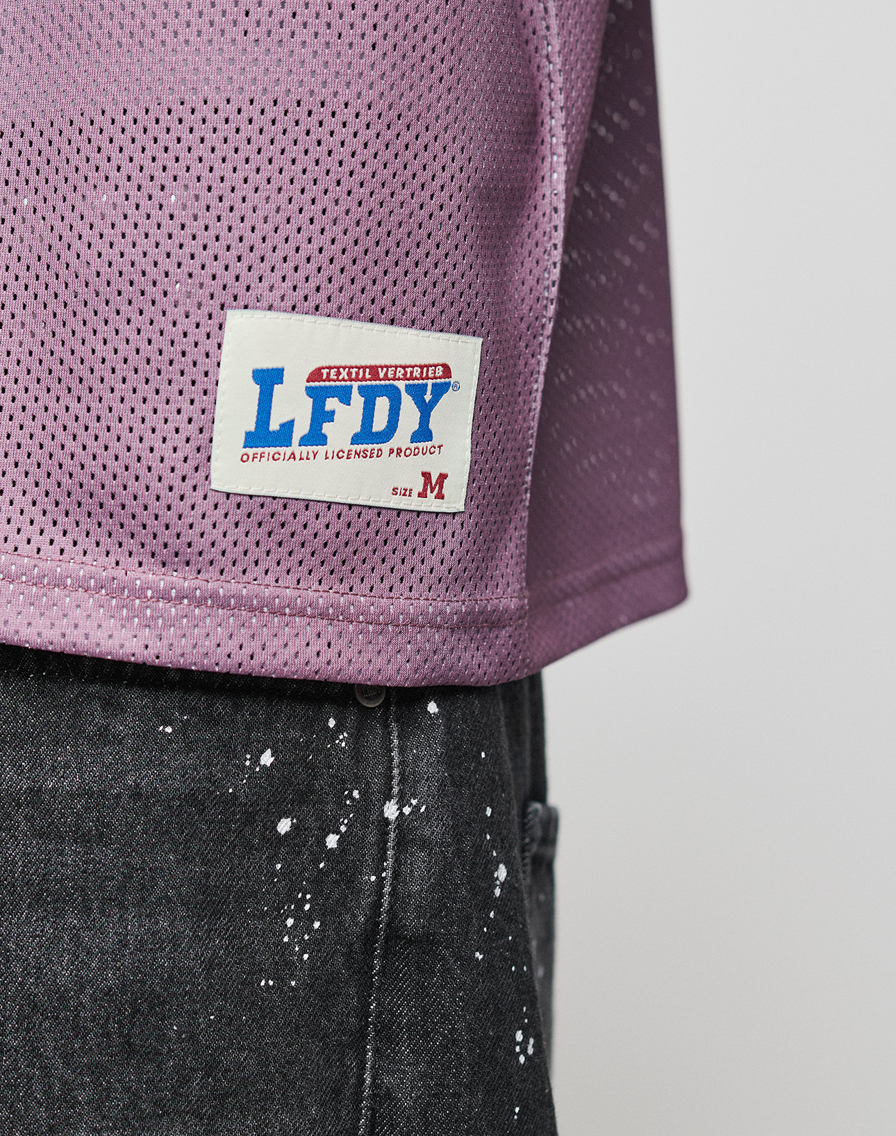 A close-up of a person wearing the LFDY Flash Jersey by LIVE FAST DIE YOUNG in oversize purple mesh, featuring a white rectangular patch labeled LFDY and OFFICIALLY LICENSED PRODUCT above black jeans with white paint splatters. Size M.