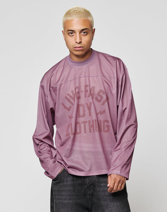 A short-haired blond person wears the LFDY Flash Jersey by LIVE FAST DIE YOUNG—an oversized mauve mesh top with a bold front print—standing with one hand in their pocket against a plain light gray background.