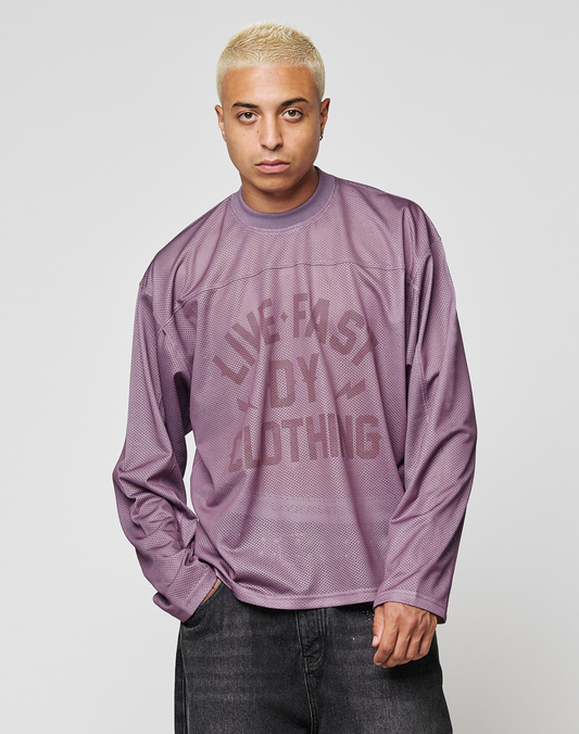 A person with short blond hair wears an oversize, long-sleeve LFDY Flash Jersey by LIVE FAST DIE YOUNG in purple mesh with a faded front print and dark pants, posing with one hand in their pocket against a plain background.