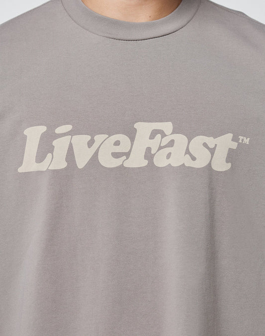 A close-up of a person wearing the LIVE FAST DIE YOUNG Basic Tee, a gray T-shirt with vintage wash and LiveFast™ printed in bold, beige letters across the chest.