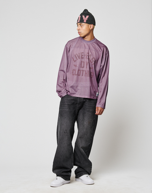 A person in an oversized purple LIVE FAST DIE YOUNG LFDY Flash Jersey, black baggy jeans, white sneakers, and a black beanie with pink letters stands against a plain light gray background, looking to the side.