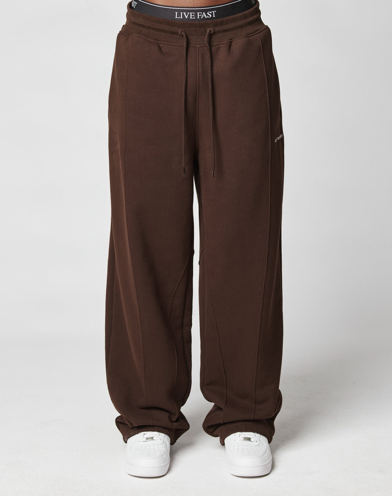A person wears wide fit, loose-fitting brown LFDY Pleated Jogger sweatpants by LIVE FAST DIE YOUNG with an elastic waistband, white sneakers, and black LIVE FAST DIE YOUNG underwear, standing against a plain white background.