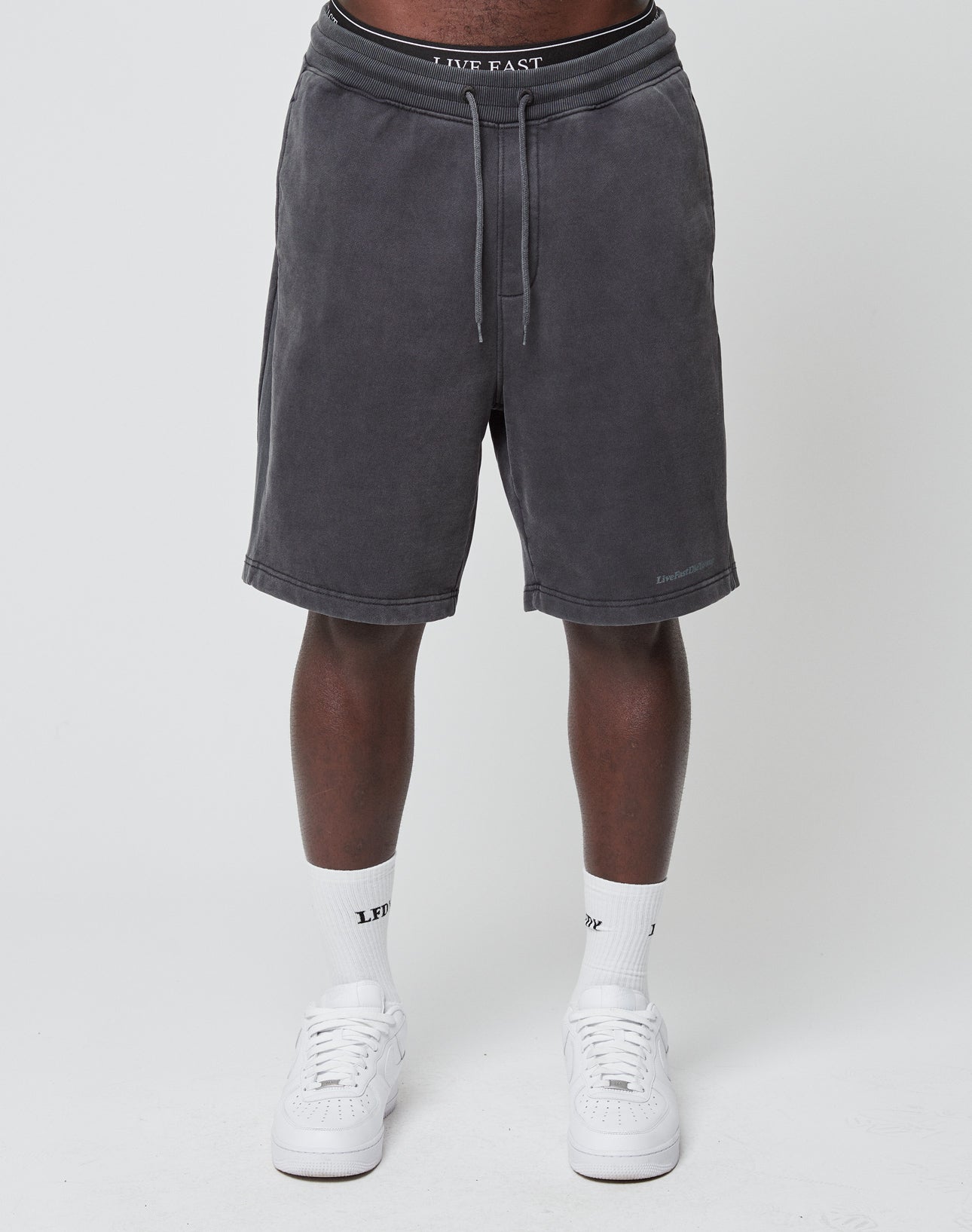 A person in LIVE FAST DIE YOUNG Basic Heavy Shorts (dark gray, vintage-washed with drawstrings), white LVFT socks, and white sneakers stands against a plain white background. Only the lower half is shown.