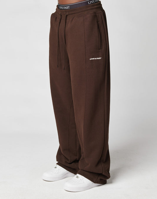 Someone wearing LIVE FAST DIE YOUNG LFDY Pleated Jogger wide fit brown sweatpants with LIVE FAST text on the left thigh, elastic waistband, and white sneakers, stands against a plain light background.| model