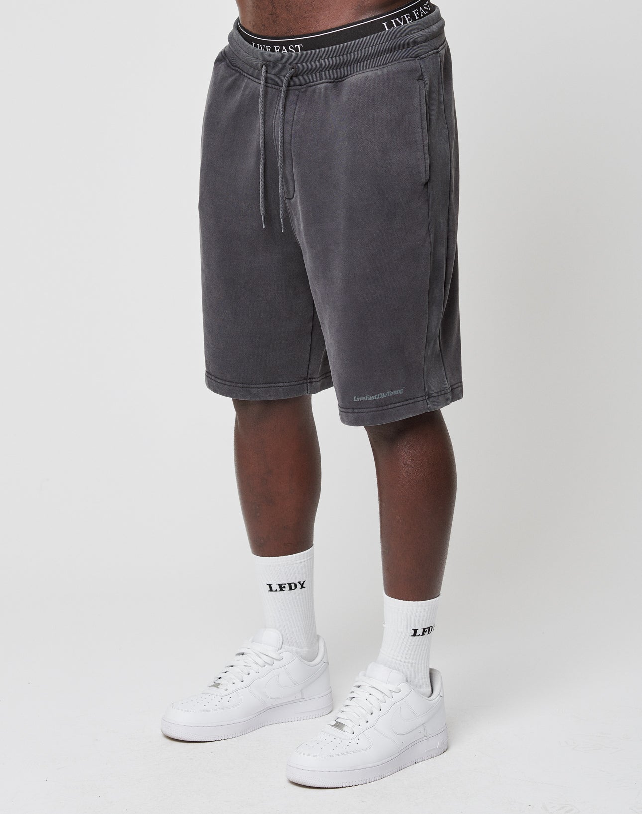 A person models the LIVE FAST DIE YOUNG Basic Heavy Shorts in charcoal gray, paired with white LFDF text socks and white sneakers against a plain white background; their upper body is cropped out and hands are not visible.
