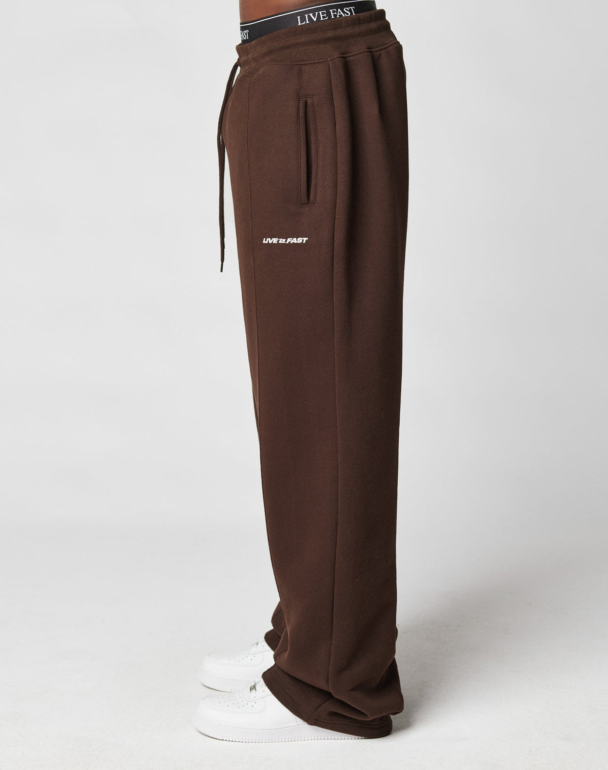 Model wears LIVE FAST DIE YOUNG’s LFDY Pleated Jogger in brown with “LIVE FAST” text near the pocket, black LIVE FAST Elastik-Bund waistband, and white sneakers. Wide fit is visible in a side profile on a plain white background.