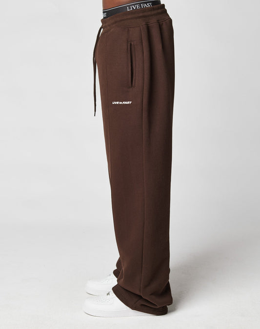 Model wears LIVE FAST DIE YOUNG’s LFDY Pleated Jogger in brown with “LIVE FAST” text near the pocket, black LIVE FAST Elastik-Bund waistband, and white sneakers. Wide fit is visible in a side profile on a plain white background.