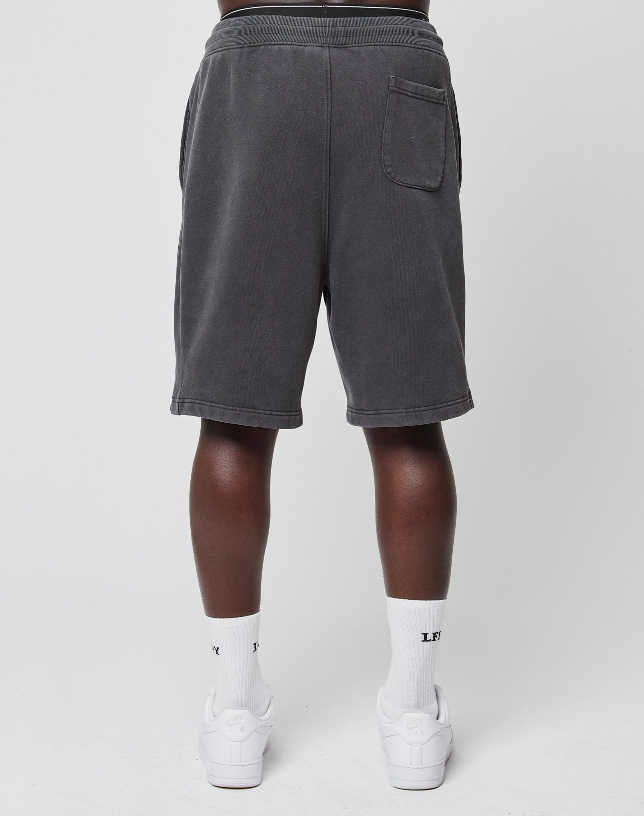 A person in LIVE FAST DIE YOUNG Basic Heavy Shorts (dark gray, Regular Fit), white socks, and white sneakers stands facing away from the camera against a plain white background.