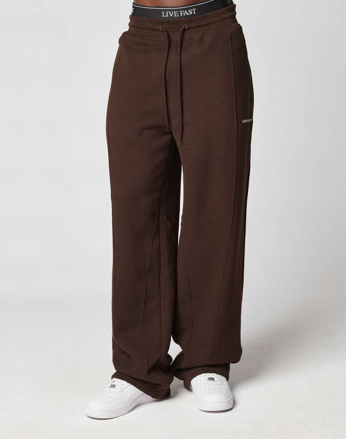 A person wearing LFDY Pleated Jogger wide-fit brown sweatpants by LIVE FAST DIE YOUNG with an elastic waistband, paired with white sneakers, stands against a plain white background.