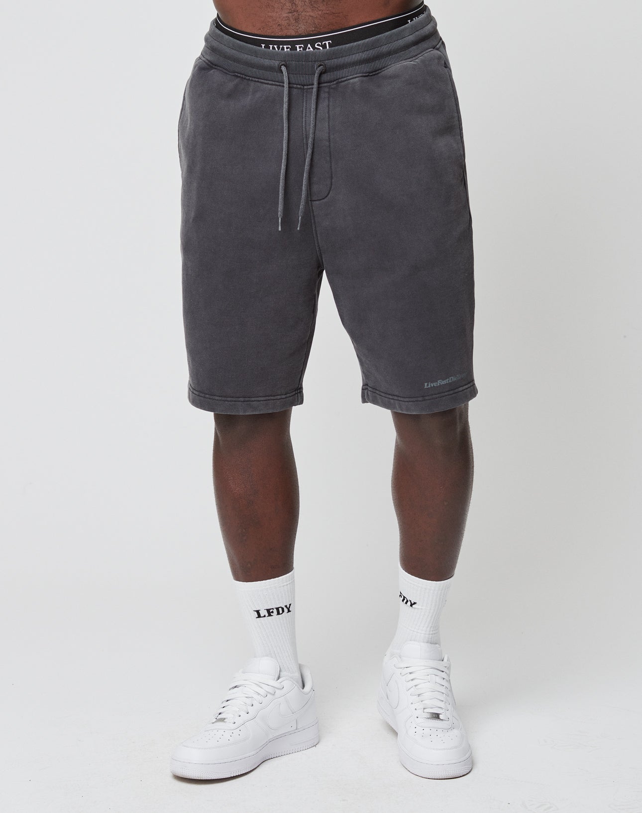 A person wearing LIVE FAST DIE YOUNG Basic Heavy Shorts in charcoal gray stands against a white background, paired with white LEDY socks and white sneakers. Only the lower half of the body is visible.