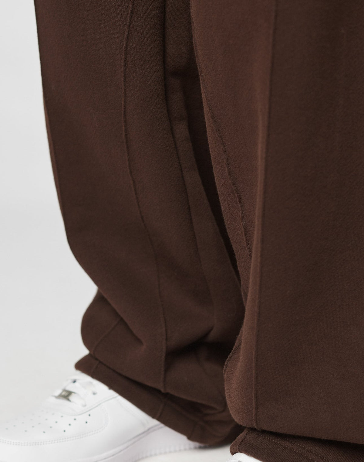 A close-up shows a person wearing LIVE FAST DIE YOUNGs LFDY Pleated Jogger—wide fit, loose brown pants with an elastic waistband—paired with white sneakers against a plain, light background. The pants just touch the shoes.