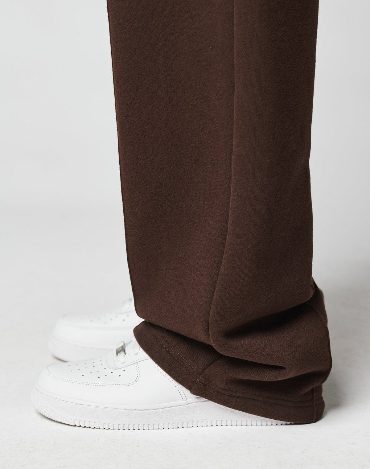 A close-up side view of a person wearing the LFDY Pleated Jogger by LIVE FAST DIE YOUNG, featuring a wide fit and Elastik-Bund in brown, draped over white perforated sneakers against a plain light backdrop.