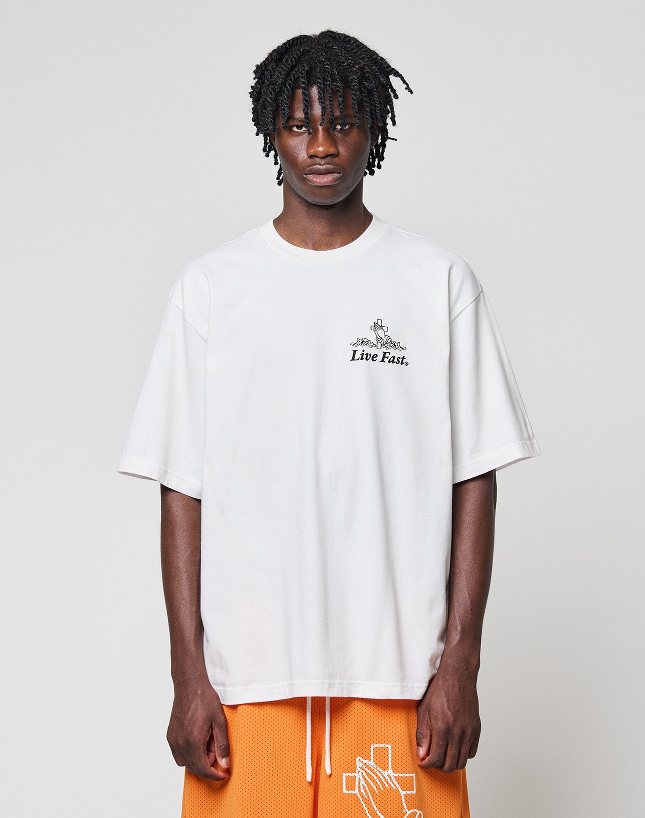 A person with medium-length braided hair wears the LFDY Praying Tee by LIVE FAST DIE YOUNG, featuring “Live Fast.” front and back prints, paired with orange basketball shorts with a white cross design, against a light gray background.