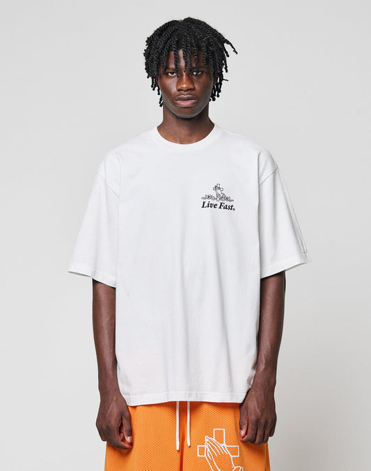 A person with medium-length braided hair wears the LFDY Praying Tee by LIVE FAST DIE YOUNG, featuring “Live Fast.” front and back prints, paired with orange basketball shorts with a white cross design, against a light gray background.