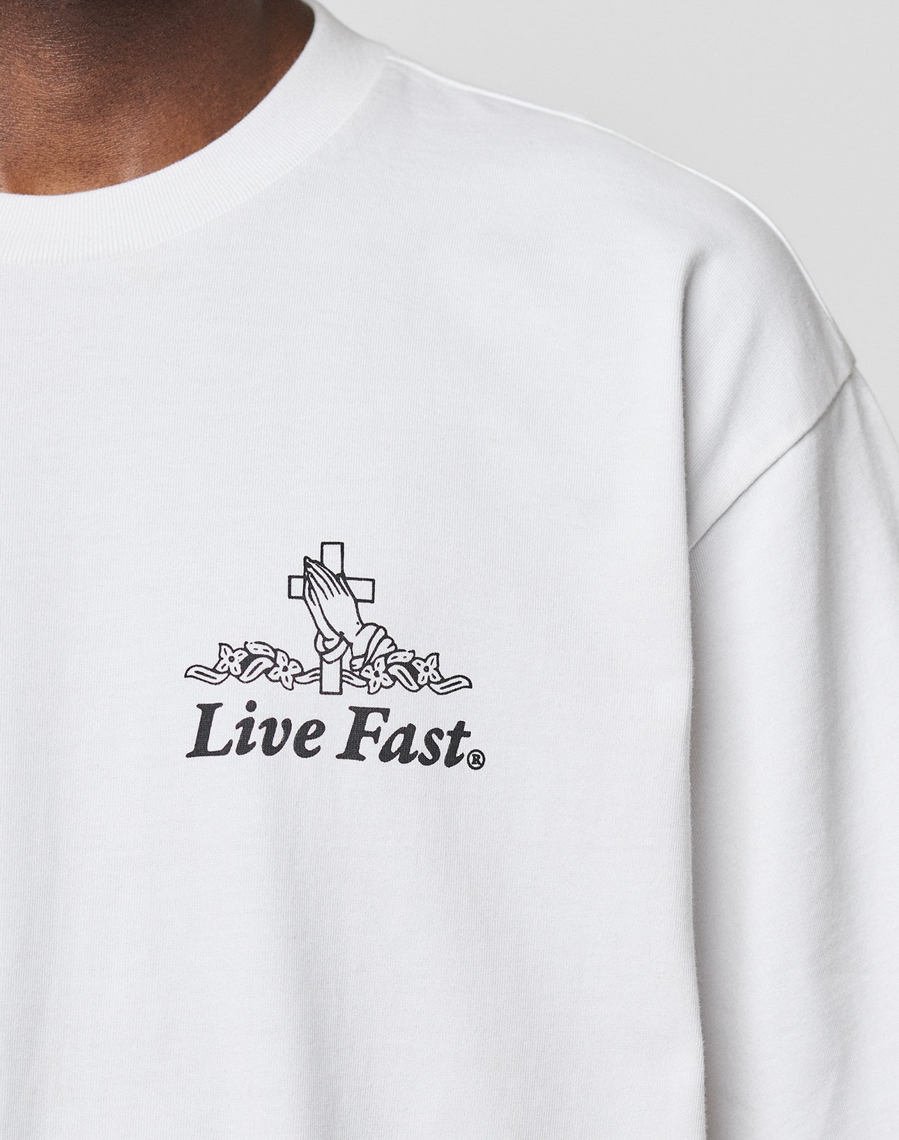 A person wears the LFDY Praying Tee by LIVE FAST DIE YOUNG, an oversized white shirt made from 100% Baumwolle with a front print of praying hands, a cross, flowers, and Live Fast below the graphic.