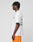 A person with short braided hair stands in profile wearing the LIVE FAST DIE YOUNG LFDY Praying Tee—an oversize white graphic t-shirt with front and back print—and orange athletic shorts against a plain light background.