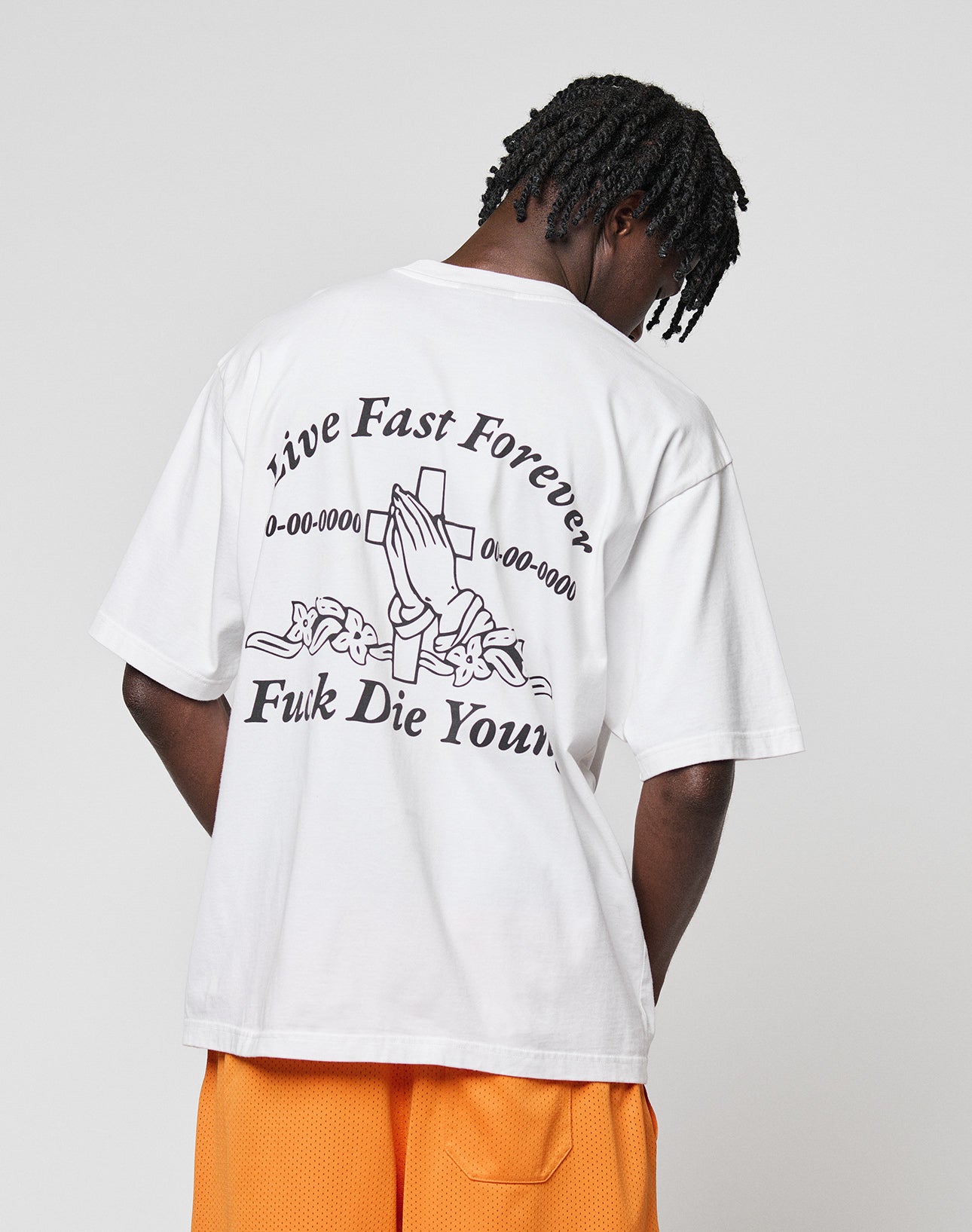 A person with short braids, facing away from the camera, wears the LIVE FAST DIE YOUNG LFDY Praying Tee—an oversize white shirt with bold front and back graphics including praying hands—and orange shorts.