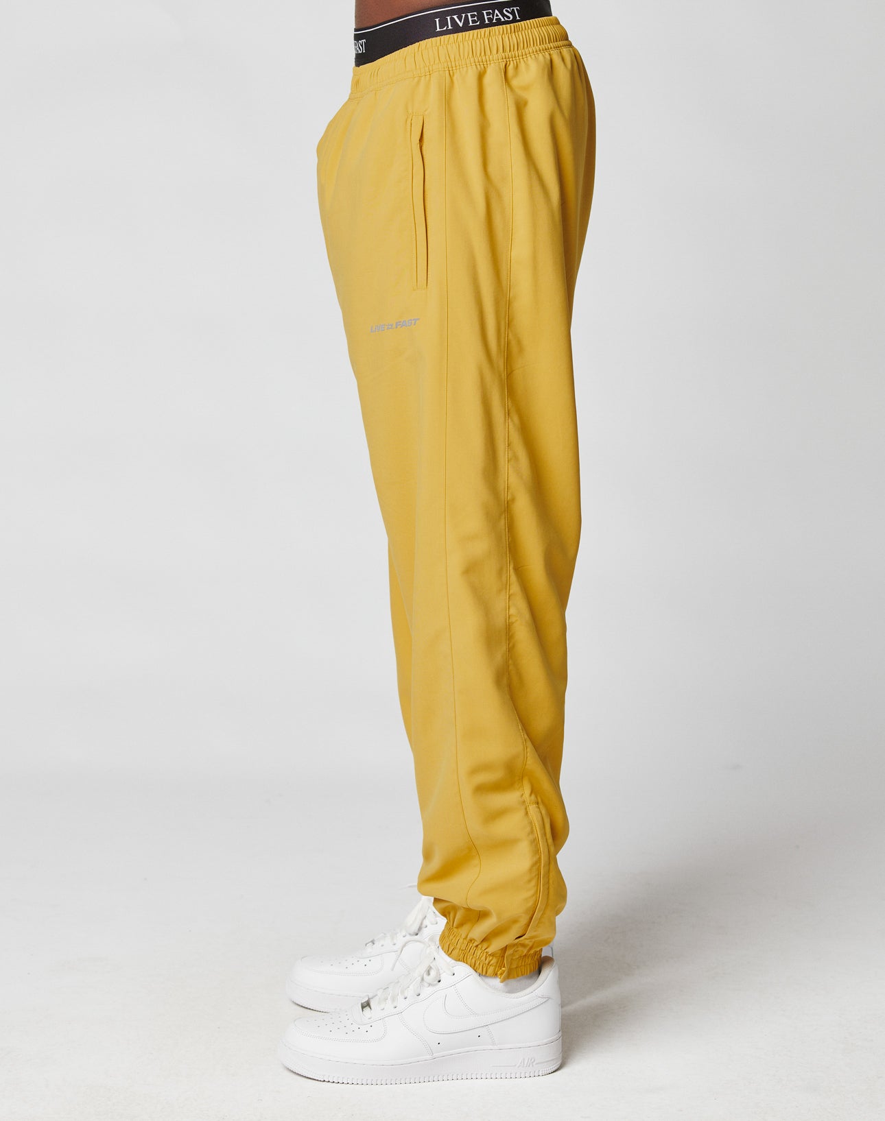 A person in LIVE FAST DIE YOUNG LFDY Trackpants—mustard yellow with Elastikbund and reflective Live Fast print—stands sideways in white sneakers against a plain white background, showing only the lower body.