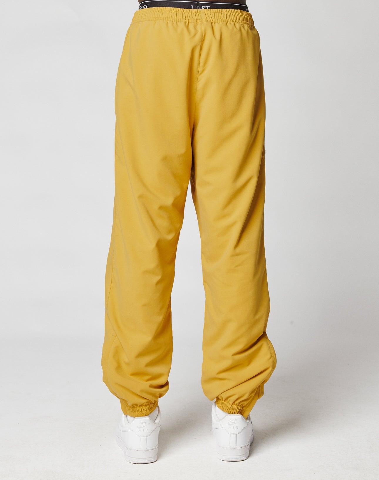 Model wearing LIVE FAST DIE YOUNG LFDY Trackpants in mustard yellow with elastic waistband and cuffs, paired with white sneakers. Photo shows the loose polyester fit from the back against a plain white background.