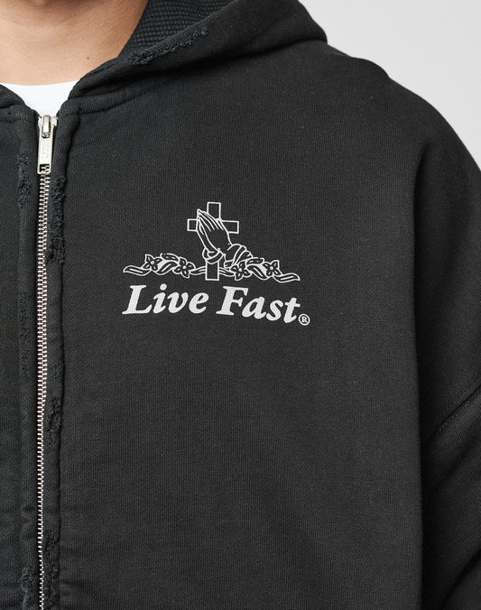 A person wears the LFDY Praying Hooded Zip by LIVE FAST DIE YOUNG, an oversized black zip-up hoodie with a white graphic of praying hands, a cross, and Live Fast on the chest. Features subtle distress-effect details for an edgy look.