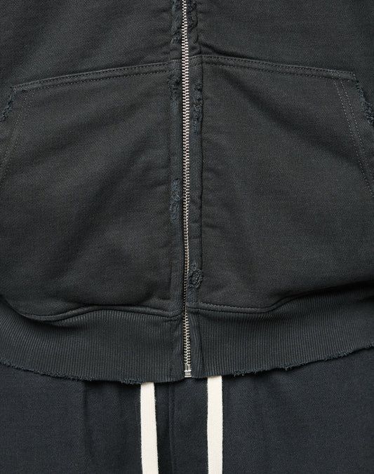 Close-up of the LFDY Praying Hooded Zip by LIVE FAST DIE YOUNG, a black oversized hoodie with a metal zipper, front pouch pocket, frayed hem Distress-Effekt, paired with dark pants featuring two vertical white stripes.