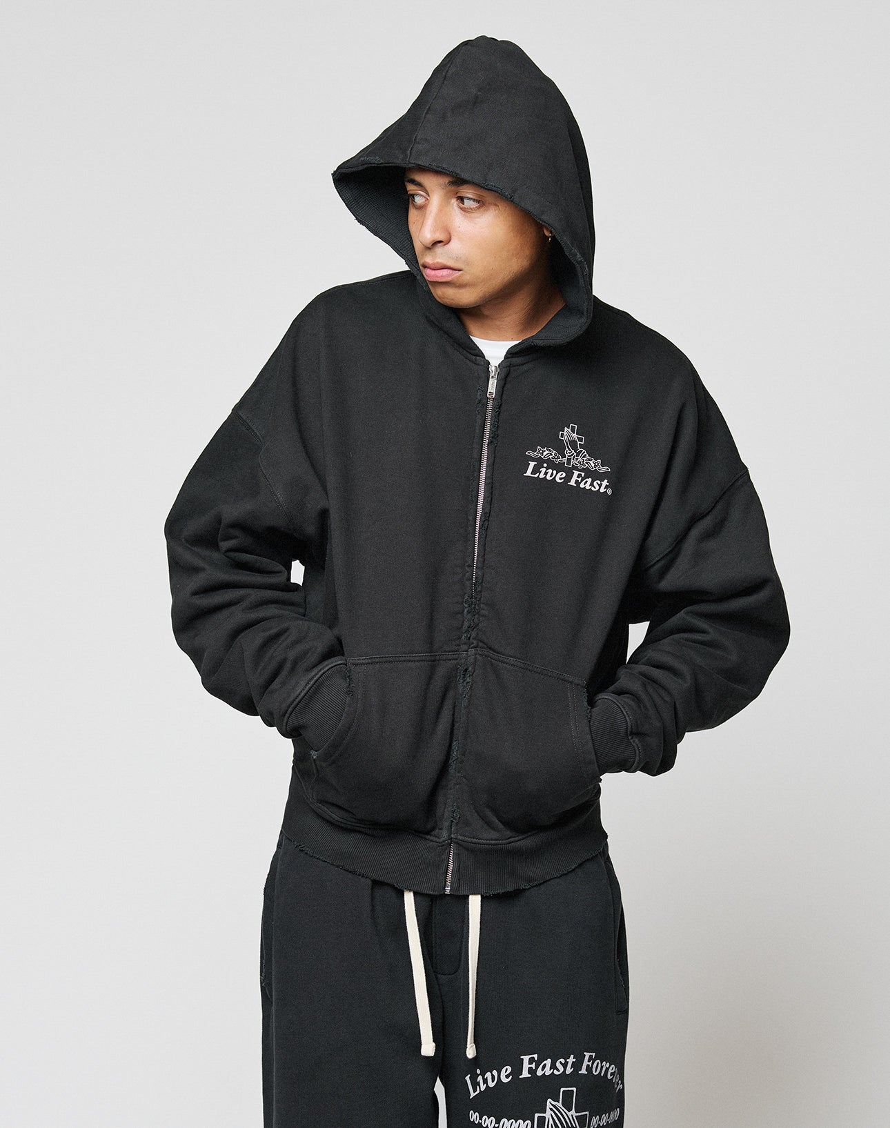A person wearing the LIVE FAST DIE YOUNG LFDY Praying Hooded Zip and matching black sweatpants with white “Live Fast” text stands against a light gray background, looking to their left.