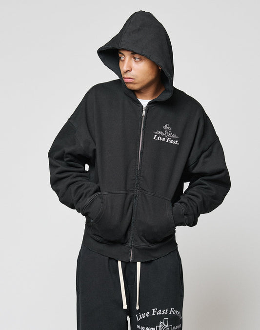 A person wearing the LIVE FAST DIE YOUNG LFDY Praying Hooded Zip and matching black sweatpants with white “Live Fast” text stands against a light gray background, looking to their left.