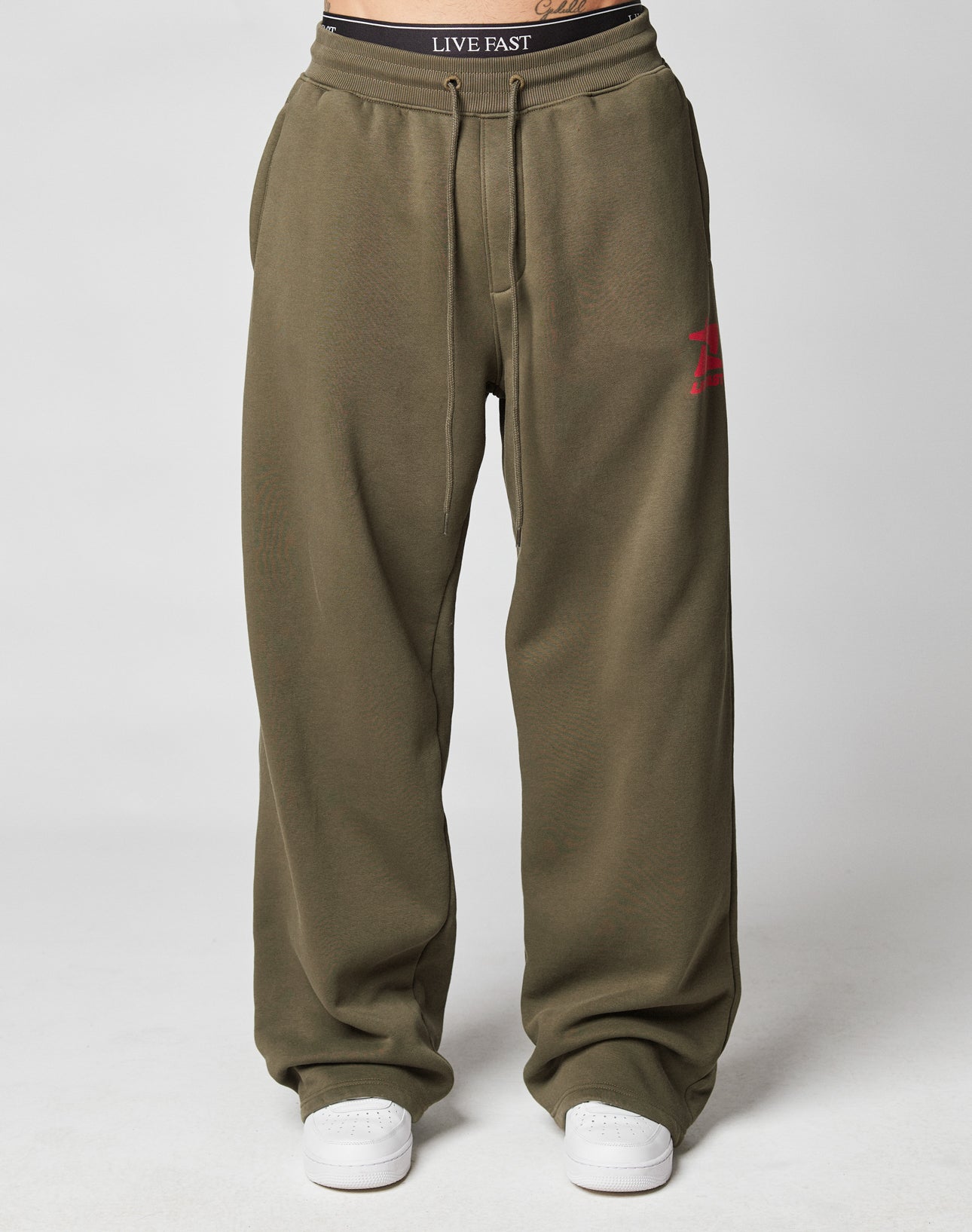 A person wears the LIVE FAST DIE YOUNG Live Fast Sweatsuit Jogger in loose-fitting olive green with an oversize fit, wide elastic waistband, drawstring waist, red A logo on one leg, white sneakers, and a black LIVE FAST waistband. Plain light background.