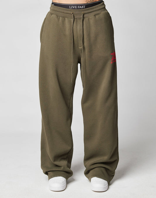 A person wears the LIVE FAST DIE YOUNG Live Fast Sweatsuit Jogger in loose-fitting olive green with an oversize fit, wide elastic waistband, drawstring waist, red A logo on one leg, white sneakers, and a black LIVE FAST waistband. Plain light background.