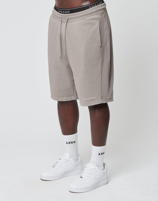 A person wearing beige Basic Heavy Shorts by LIVE FAST DIE YOUNG with subtle Vintage-Waschung, white LEFY socks, and white sneakers stands against a plain white background. The waistband of the shorts shows LIVE FAST. Face is not visible.